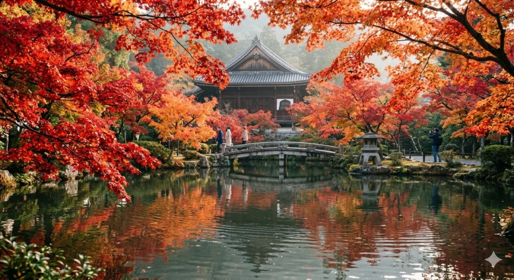 A serene autumn landscape photograph looking across a reflective pond at the Eikan-do Zenrin-ji Temple in Kyoto, Japan, during peak fall foliage in mid-November. Bright crimson and fiery orange maple trees frame the traditional dark wood architecture and a curved stone bridge, casting colorful reflections in the still water.